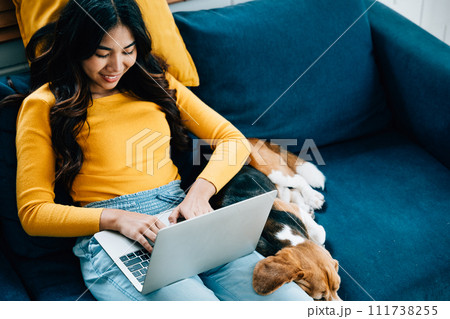 In her home office on the sofa, a woman works diligently on her laptop while her Beagle dog enjoys a nap. Their friendship and smiles define this cozy workspace. Pet love 111738255