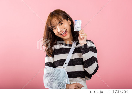 Smiling Asian woman with a broken arm wears an arm splint and handles emergency medical bills using a credit card. Happy female with a sling-supported hand in a studio shot on pink background. 111738256