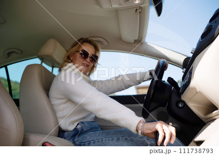 A blonde woman in a white sweater and jeans is driving. Happy woman sitting in a car with a white interior. A blonde woman in a white sweater and jeans is driving. Happy woman sitting in a car with a white interior. 111738793