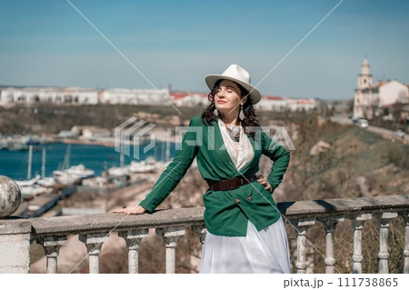 Woman walks around the city, lifestyle. Happy woman in a green jacket, white skirt and hat is sitting on a white fence with balusters overlooking the sea bay and the city. 111738865
