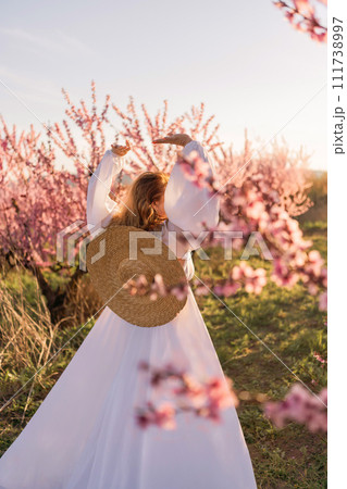 Woman blooming peach orchard. Against the backdrop of a picturesque peach orchard, a woman in a long white dress and hat enjoys a peaceful walk in the park, surrounded by the beauty of nature. Woman blooming peach orchard. Against the backdrop of a picturesque peach orchard, a woman in a long white dress and hat enjoys a peaceful walk in the park, surrounded by the beauty of nature. 111738997