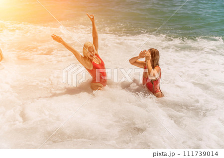 Women ocean play. Seaside, beach daytime, enjoying beach fun. Two women in red swimsuits enjoying themselves in the ocean waves and raising their hands up. 111739014