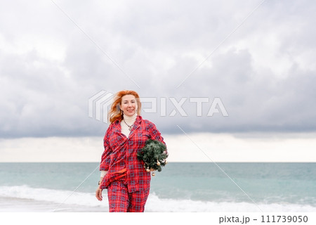 Sea Lady in plaid shirt with a christmas tree in her hands enjoys beach. Coastal area. Christmas, New Year holidays concep 111739050