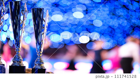 Close-up of golden trophies lined up at a dance event. Collection of trophies. Award cups closeup with space for text on neon background Close-up of golden trophies lined up at a dance event. Collection of trophies. Award cups closeup with space for text on neon background 111741528