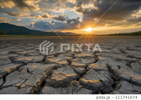 Sunbeams pierce through clouds over a parched earth, where deep cracks in the dry soil create a dramatic testament to the severity of drought conditions against a backdrop of distant mountains. 111741674