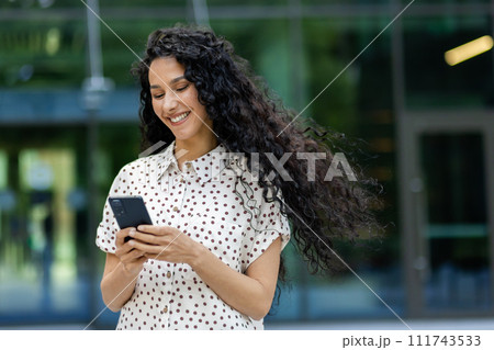 Young beautiful Latin American woman walking in the city with phone in hands, business woman after working day, smiling with satisfaction, student using application on smartphone. Young beautiful Latin American woman walking in the city with phone in hands, business woman after working day, smiling with satisfaction, student using application on smartphone. 111743533
