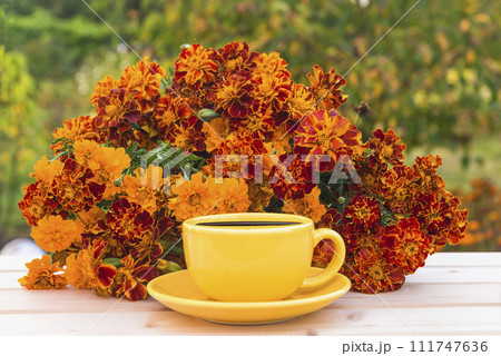 Summer leisure activities concept; yellow cup of coffee and bunch of orange marigold flowers on a wooden table in green garden 111747636