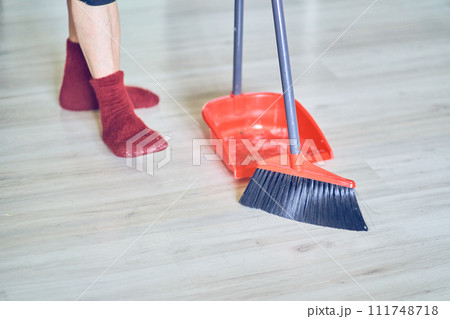 Close up view of a red broom and plastic dustpan on the floor with person's legs in red socks. Worker, cleaner using cleaning supplies with copy space. Concept of cleaning home, dust, sweeping floor. 111748718