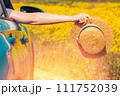 Feminine hand holding straw hat from the opened window of the car in the meadow of yellow flowers 111752039