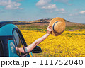 Feminine hand holding straw hat from the opened window of the car, among the yellow wild flowers. 111752040