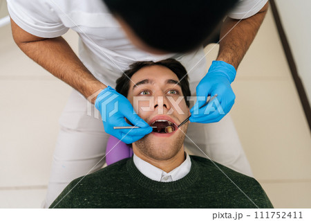 Portrait of handsome male patient lying in stomatology seat with open mouth during unrecognizable professional dentist examining teeth, with dental tool in dentistry clinic. Concept of stomatology. 111752411