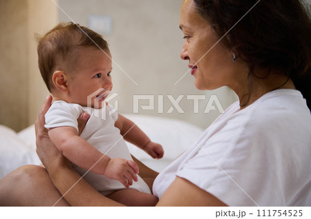 Loving mom sitting on the bed, holding her baby boy, communicating and smiling to him, establishing emotional contact 111754525