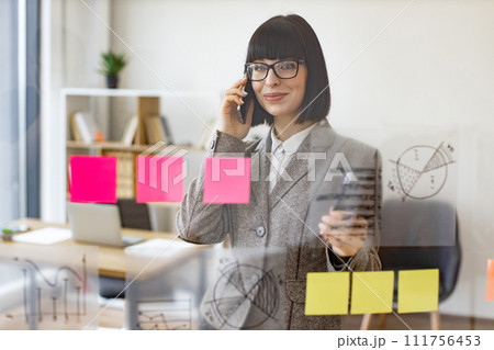 Businesswoman talking on phone while analyzing growth of charts on glass board. Businesswoman talking on phone while analyzing growth of charts on glass board. 111756453