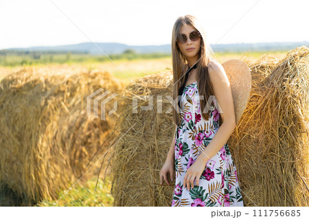 beautiful glamorous girl in a dress and a straw hat on a field near a haystack. 111756685