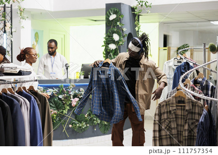 Shopper enjoying music in headphones while browsing for trendy clothes in mall. Showroom customer in earphones holding casual plaid shirt on hanger to try on in fitting room Shopper enjoying music in headphones while browsing for trendy clothes in mall. Showroom customer in earphones holding casual plaid shirt on hanger to try on in fitting room 111757354