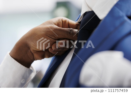 African american man arms fixing tie on male mannequin in fashion showroom. Boutique seller wearing formal suit on model and adjusting jacket, showcasing clothes to customer in mall 111757459