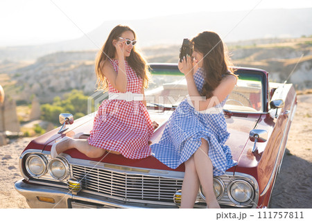 two beautiful girl in a retro dress are photographed on camera on mountain landscape in Cappadocia near a red retro car two beautiful girl in a retro dress are photographed on camera on mountain landscape in Cappadocia near a red retro car 111757811