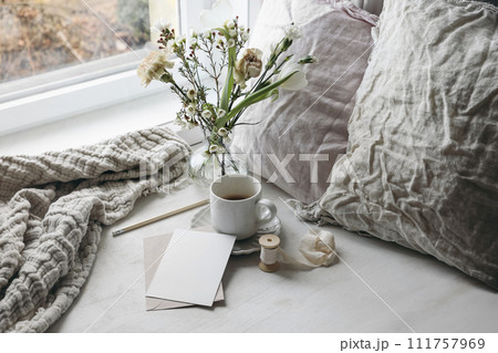 Easter spring still life. Cup of coffee and floral bouquet on window sill. White tulips, carnation flowers in vase. Blank greeting card mockup. Linen pillows, cushion. Scandinavian home, breakfast. Easter spring still life. Cup of coffee and floral bouquet on window sill. White tulips, carnation flowers in vase. Blank greeting card mockup. Linen pillows, cushion. Scandinavian home, breakfast. 111757969