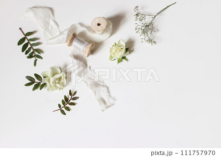 Floral wedding, birthday table composition. Gypsophila, rose flowers, green lentisk leaves and silk ribbons with wooden spools isolated on white table background. Flat lay, top view. Copy space, no Floral wedding, birthday table composition. Gypsophila, rose flowers, green lentisk leaves and silk ribbons with wooden spools isolated on white table background. Flat lay, top view. Copy space, no 111757970