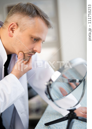 the man is a doctor, beautician looks at his skin in the mirror the man is a doctor, beautician looks at his skin in the mirror 111758253