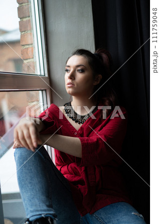 a beautiful young girl in a red shirt with a handmade jewelry necklace around her neck is posing near the window 111759048