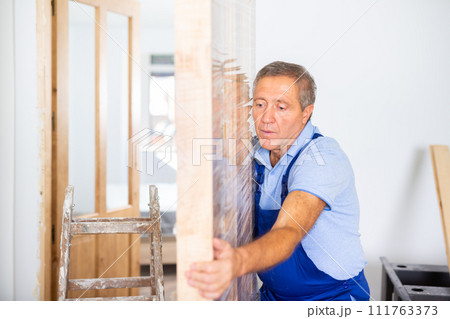 Professional male craftsman in overalls dragging the door construction into an apartment Professional male craftsman in overalls dragging the door construction into an apartment 111763373