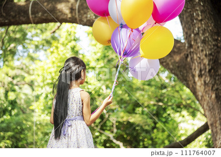 Cheerful cute girl holding balloons running on green meadow white cloud and blue sky with happiness. Hands holding vibrant air balloons play on birthday party happy times summer on sunlight outdoor 111764007