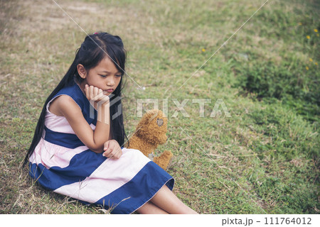 Sad girl hugging teddy bear sadness alone in green garden park. Lonely girl feeling sad unhappy sitting outdoors with best friend toy. Autism child play teddy bear best friend. Family violence concept Sad girl hugging teddy bear sadness alone in green garden park. Lonely girl feeling sad unhappy sitting outdoors with best friend toy. Autism child play teddy bear best friend. Family violence concept 111764012