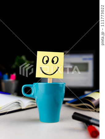 Smiling face drawn on sticky note inside a cup of coffee at the office desk. Every morning wake up with the smile Smiling face drawn on sticky note inside a cup of coffee at the office desk. Every morning wake up with the smile 111773022