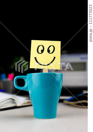 Smiling face drawn on sticky note inside a cup of coffee at the office desk. Every morning wake up with the smile Smiling face drawn on sticky note inside a cup of coffee at the office desk. Every morning wake up with the smile 111773023