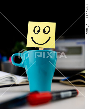 Smiling face drawn on sticky note inside a cup of coffee at the office desk. Every morning wake up with the smile Smiling face drawn on sticky note inside a cup of coffee at the office desk. Every morning wake up with the smile 111773024