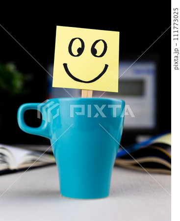 Smiling face drawn on sticky note inside a cup of coffee at the office desk. Every morning wake up with the smile Smiling face drawn on sticky note inside a cup of coffee at the office desk. Every morning wake up with the smile 111773025