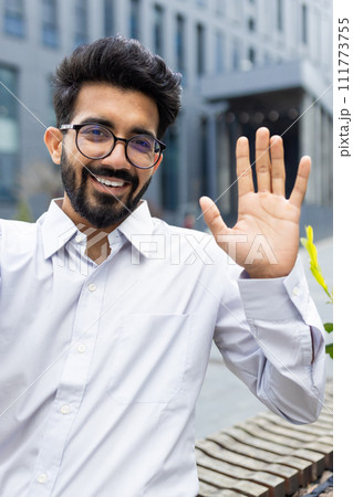 Smiling modern businessman waving hello, showing a friendly gesture, standing outdoors with a cityscape backdrop. 111773755