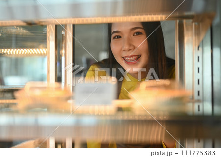 Smiling female small business owner arranging cakes in showcase display in her bakery shop 111775383