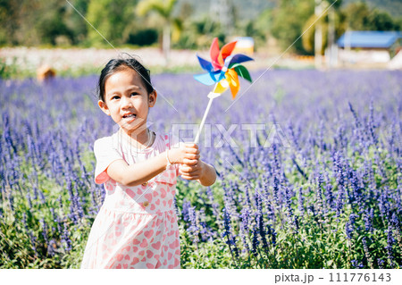 A cheerful little girl stands in a flower garden holding a toy pinwheel. The vibrant springtime evokes joy and flying pinwheels embodying childhood happiness and freedom in nature's sunny embrace. 111776143