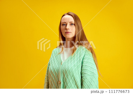 Portrait of one calm young and beautiful woman with red hair and freckles looking at camera against yellow studio background. Concept of human emotions. 111776673