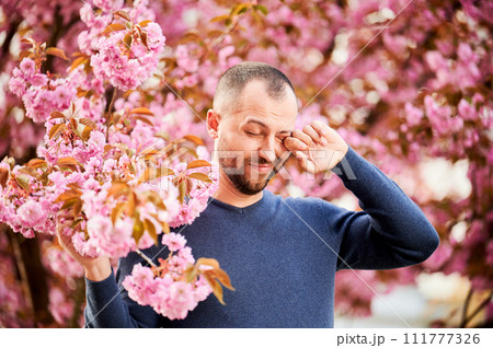 Man allergic suffering from seasonal allergy at spring in blossoming garden at springtime. Bearded man feeling itchy eyes in front of blooming tree. Spring allergy concept. 111777326