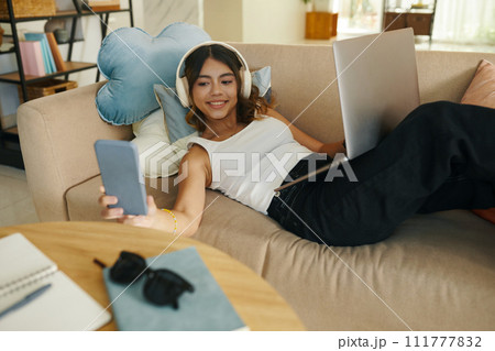 Smiling girl checking notification on phone when lying on couch and working on laptop Smiling girl checking notification on phone when lying on couch and working on laptop 111777832