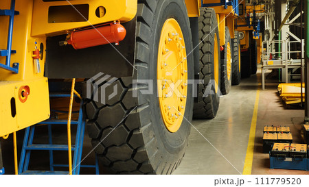 Assembly line of large dump trucks. Finished dump trucks are lined up in a production shop. A close-up of the machine wheels. Production of large yellow dump trucks. 111779520