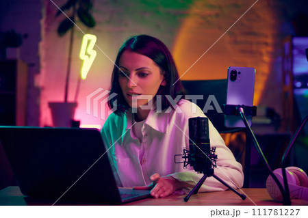 Young woman, blogger sitting in modern room with neon lights, working with laptop, leading online video lesson. Online education Young woman, blogger sitting in modern room with neon lights, working with laptop, leading online video lesson. Online education 111781227