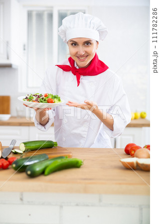 Smiling female chef in white uniform offering plate of vegetable salad in home kitche Smiling female chef in white uniform offering plate of vegetable salad in home kitche 111782286
