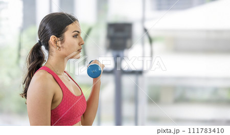 Young Focused female athlete performing a bicep curl with a blue dumbbell in a bright gym. Young Focused female athlete performing a bicep curl with a blue dumbbell in a bright gym. 111783410