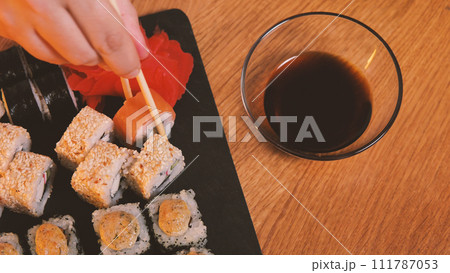 A set of delicious Japanese sushi rolls on a black stone board. View from above. Chopsticks are used to remove the sushi from the set. A bowl of soy sauce stands nearby. A set of delicious Japanese sushi rolls on a black stone board. View from above. Chopsticks are used to remove the sushi from the set. A bowl of soy sauce stands nearby. 111787053