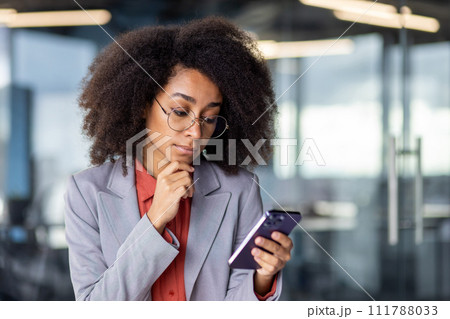 Thoughtful latino woman in grey suit holding chin with hand while looking at smartphone screen indoors. Doubting office worker getting text message with business offer and pondering about answer. Thoughtful latino woman in grey suit holding chin with hand while looking at smartphone screen indoors. Doubting office worker getting text message with business offer and pondering about answer. 111788033