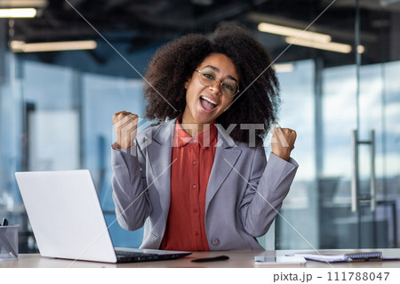 Happy woman with brunette bushy hair bending hands with clenched fists in victory gesture while sitting in office. Excited administrator celebrating finish of busy day and approximation of weekend. Happy woman with brunette bushy hair bending hands with clenched fists in victory gesture while sitting in office. Excited administrator celebrating finish of busy day and approximation of weekend. 111788047