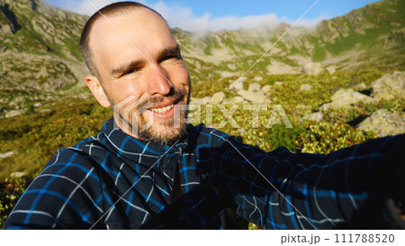 The man's face is red from tanning. A bearded Caucasian man with a short haircut is smiling as he looks into the camera. Against a background of beautiful mountains. A travelling blogger. 111788520
