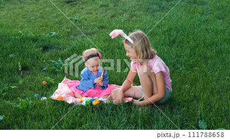 Adorable baby with older sister playing with painted eggs on a grass. Easter hunt concept 111788658