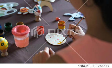 Young woman making Easter decoration at table at home, painting wooden egg Young woman making Easter decoration at table at home, painting wooden egg 111788675