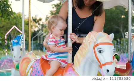 Young mother sitting her baby on a vintage horse carousel in funfair or an amusement park in the evening 111788680