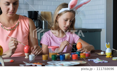 Child girl in bunny ears with her mother painting Easter eggs at table in the kitchen 111788696